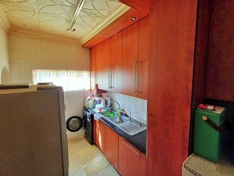 A functional scullery or laundry area featuring wood-finish cabinetry, a stainless steel sink, tiled splashbacks, and a decorative ceiling.