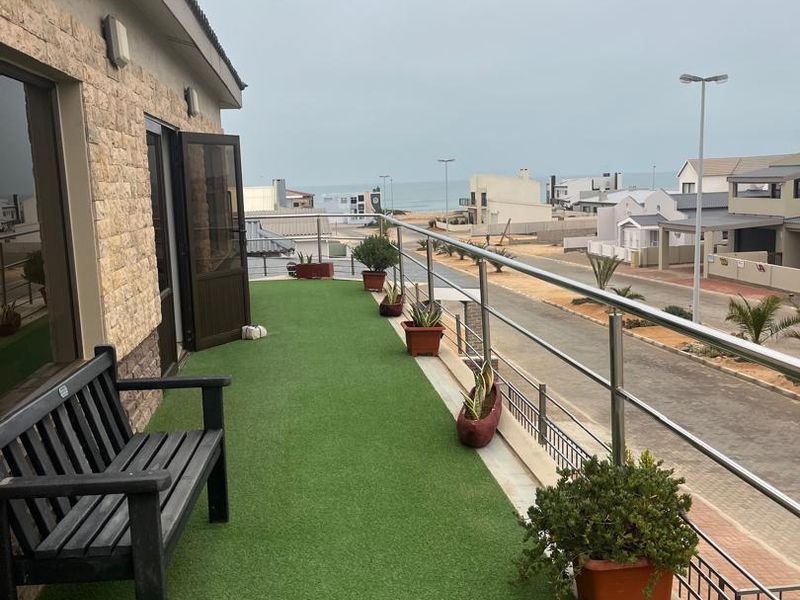 A spacious balcony featuring artificial grass flooring, a stainless steel balustrade, and stone cladding, offering a view of the surrounding neighborhood and street.