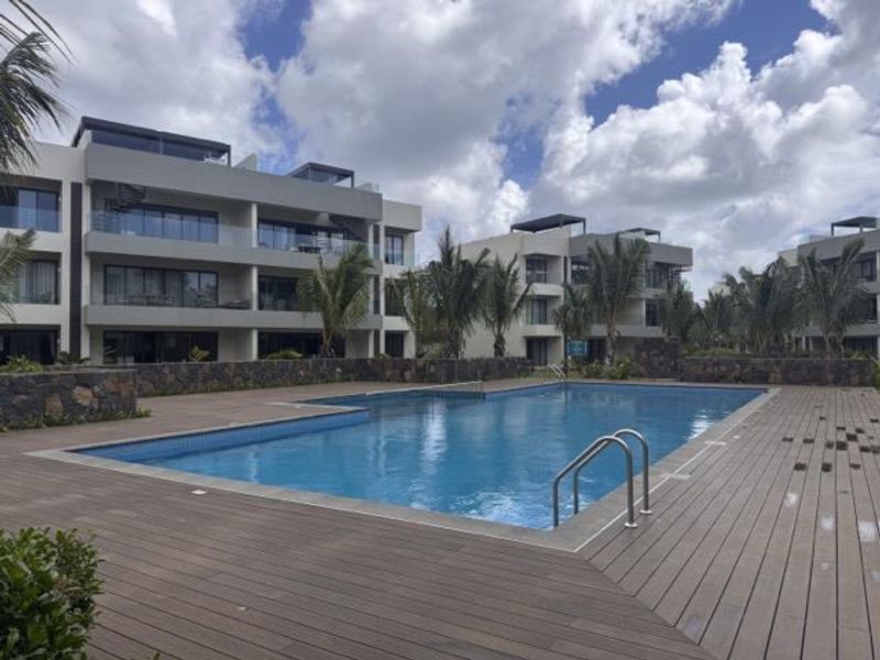 A well-maintained communal swimming pool area featuring timber decking, a stone retaining wall, and modern apartment buildings in the background.