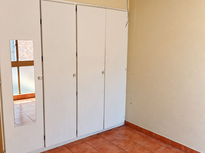 An empty bedroom featuring built-in cupboards with a mirrored panel, a ceiling fan, and terracotta-style tiled flooring.