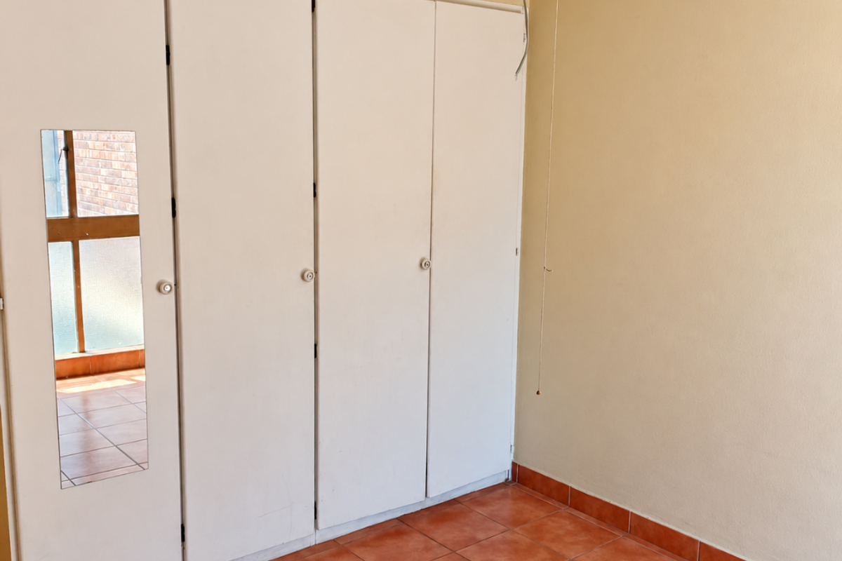 An empty bedroom featuring built-in cupboards with a mirrored panel, a ceiling fan, and terracotta-style tiled flooring.