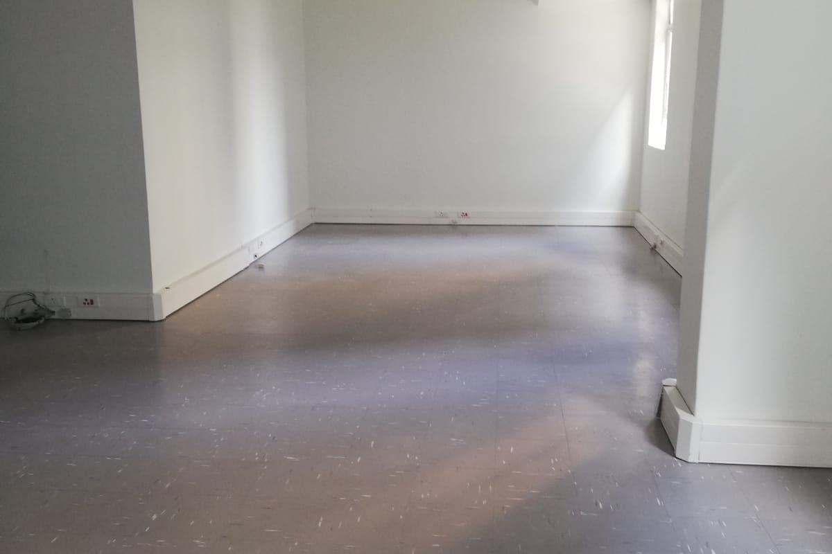 A spacious, empty office area featuring neutral white walls, durable vinyl flooring, and exposed industrial-style ceiling cable trays.