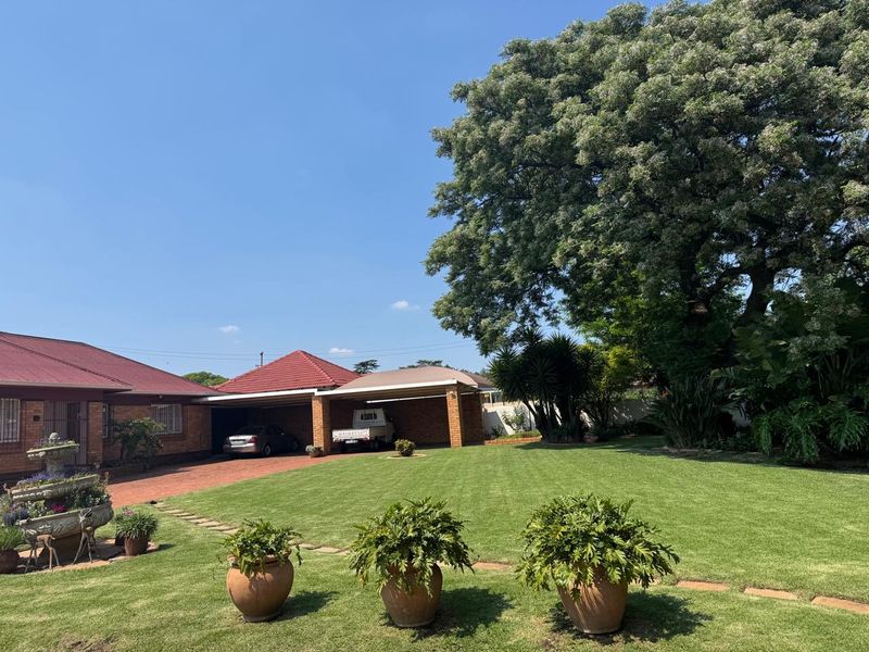 Spacious rear garden featuring a well-maintained lawn, mature trees, and a double brick carport with a paved driveway.