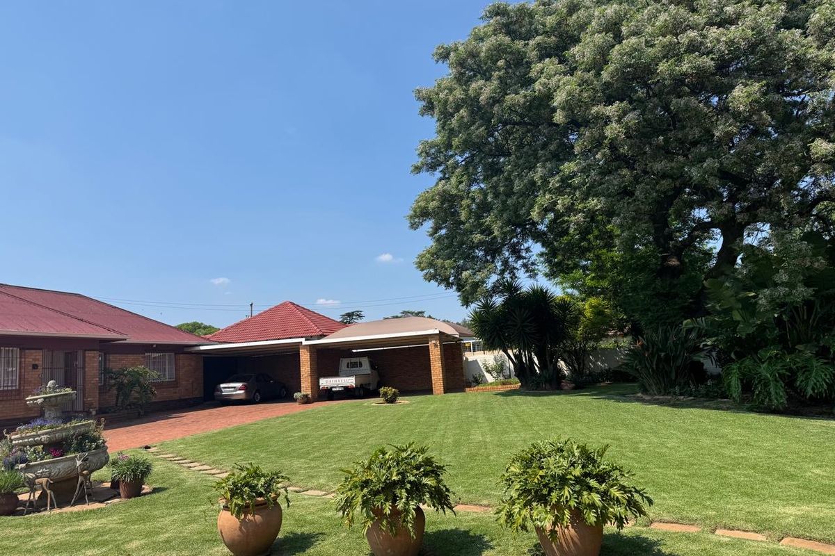 Spacious rear garden featuring a well-maintained lawn, mature trees, and a double brick carport with a paved driveway.
