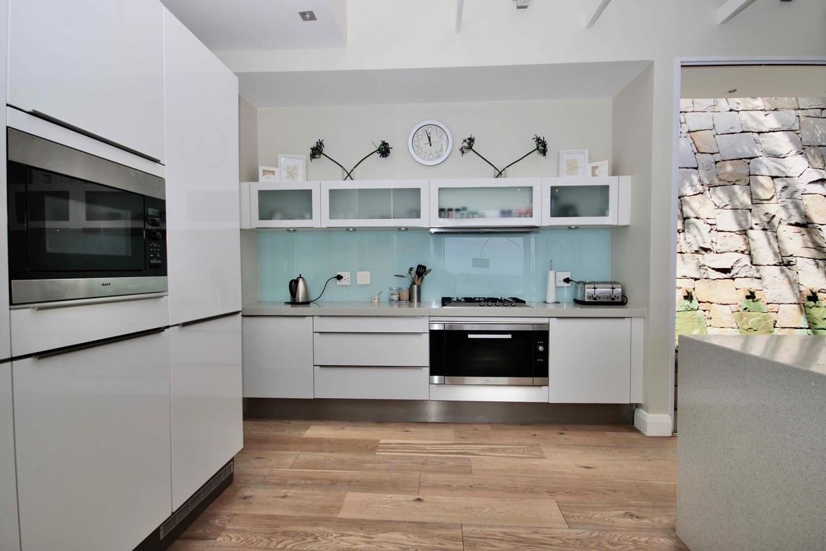 Modern kitchen featuring sleek white cabinetry, integrated oven and microwave, gas hob, and a light blue glass splashback with timber-look flooring.