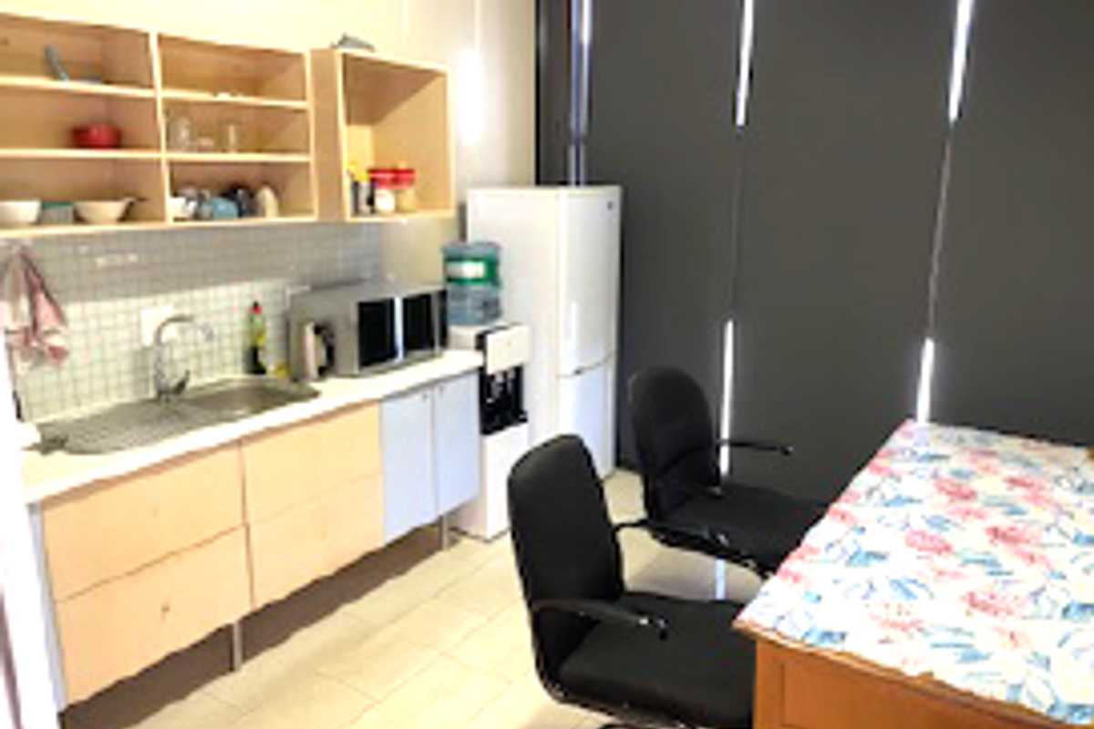 A functional office kitchenette featuring light wood cabinetry, a stainless steel sink, tiled backsplash, and roller blinds.