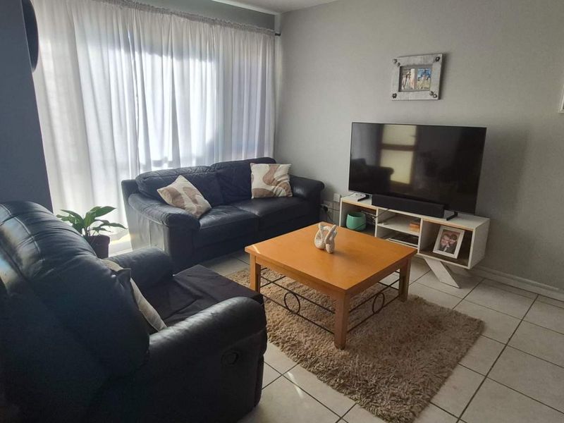 A functional living room featuring neutral tiled flooring, large windows with venetian blinds, and sheer curtains, providing a bright and comfortable living space.