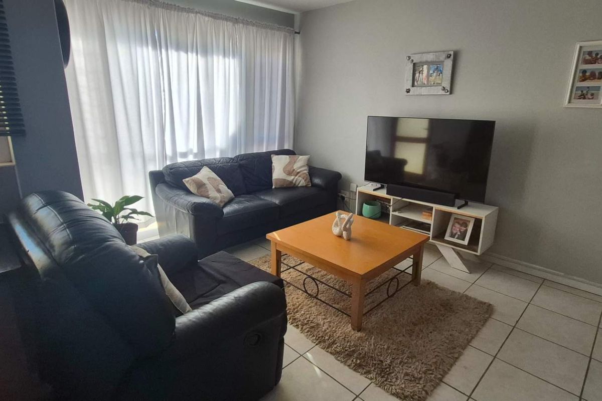 A functional living room featuring neutral tiled flooring, large windows with venetian blinds, and sheer curtains, providing a bright and comfortable living space.