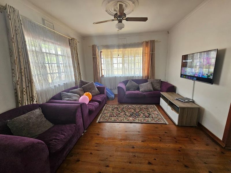 A living room featuring polished hardwood flooring, a ceiling fan with a decorative ceiling rose, and dual windows with curtain rails.