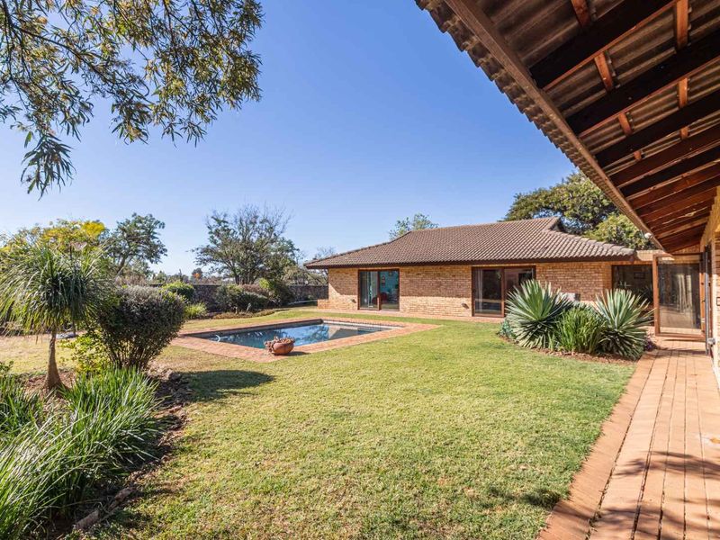 A spacious rear garden featuring a swimming pool, brick patio, and covered walkway, set against a classic brick house with exposed timber roof beams.