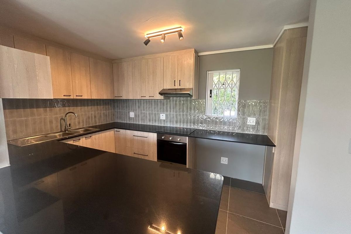 Modern kitchen featuring light wood cabinetry, dark granite countertops, integrated oven, extractor fan, and a tiled backsplash with a secure window.