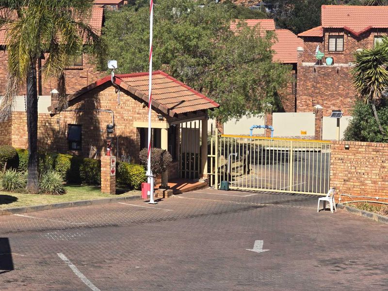 View of the complex entrance featuring a brick guardhouse, security boom gate, and metal access gate with paved driveway.