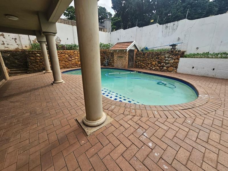 Outdoor pool area featuring a covered patio with concrete columns, brick paving, and a stone retaining wall, enclosed by a perimeter wall with electric fencing.