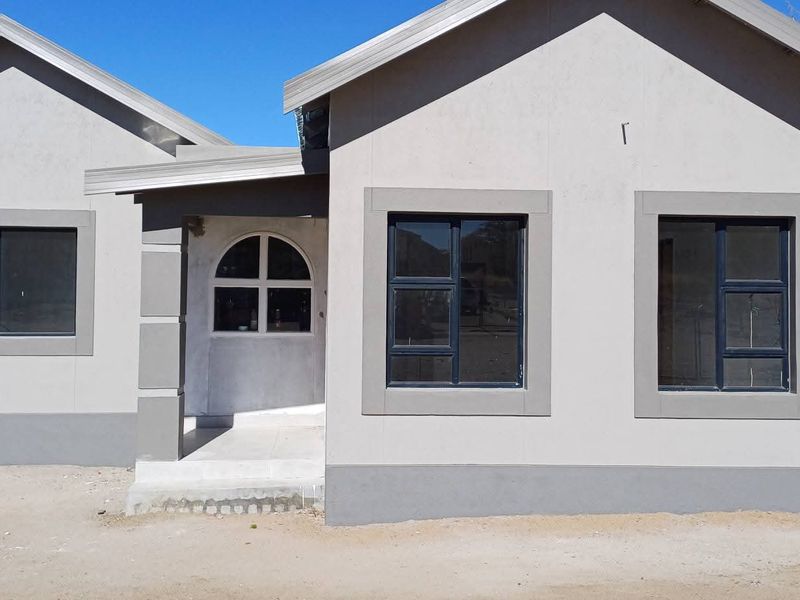 A modern, single-story residential house featuring a clean plastered facade, dark aluminium window frames, and a covered entrance porch.
