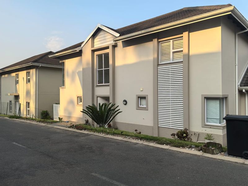 A well-maintained townhouse exterior featuring a neutral plastered facade, tiled roof, and modern louvre shutters, situated along a paved residential street.