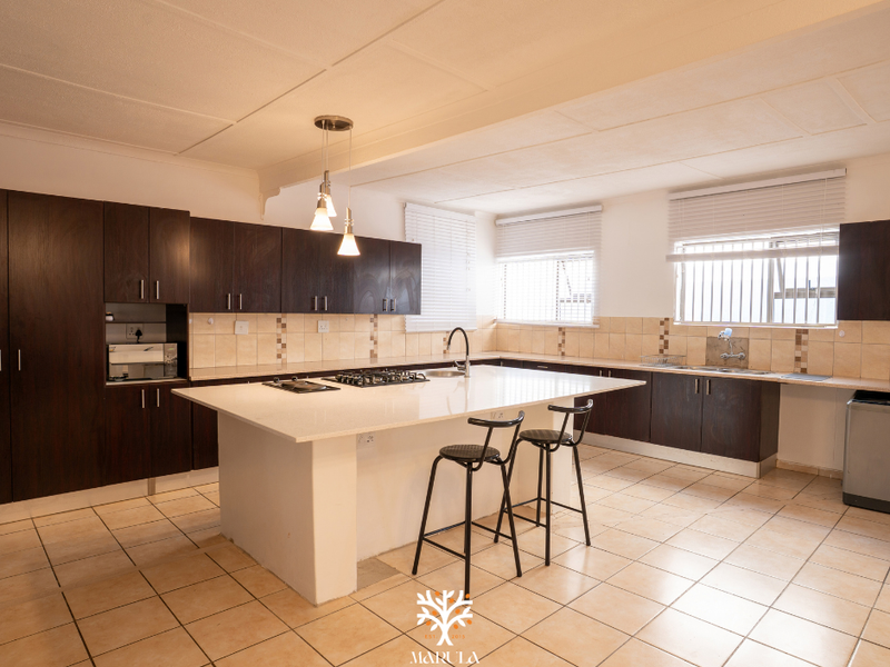 Spacious kitchen featuring a large central island with a gas hob, dark wood cabinetry, stone countertops, and tiled flooring throughout.