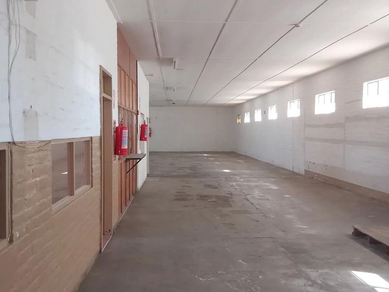 A spacious, empty industrial utility area featuring concrete flooring, exposed brick walls, and multiple windows providing natural light.