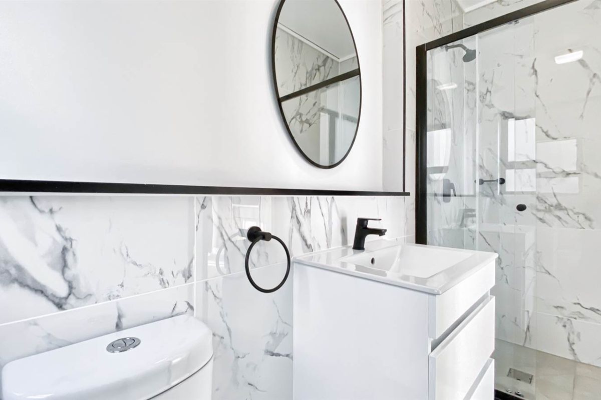 Modern bathroom featuring marble-look wall tiles, a sleek vanity with black fixtures, a circular mirror, and a glass-enclosed shower.