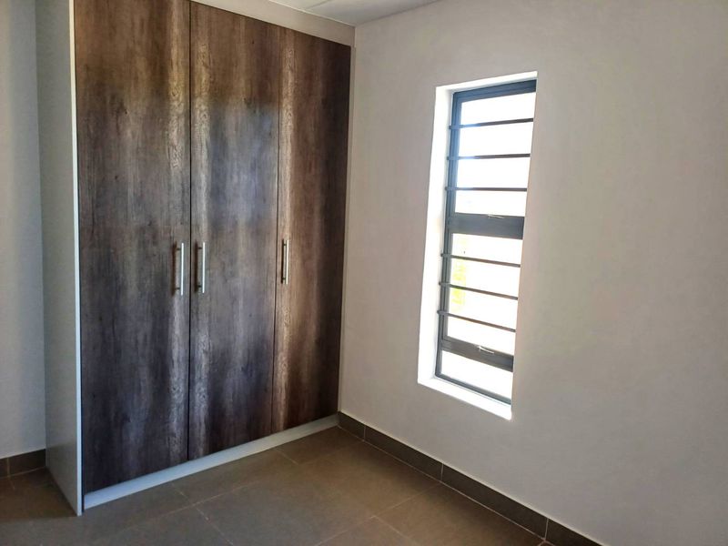 A clean, empty bedroom featuring modern wood-finish built-in cupboards, neutral tiled flooring, and a louvered window providing natural light.