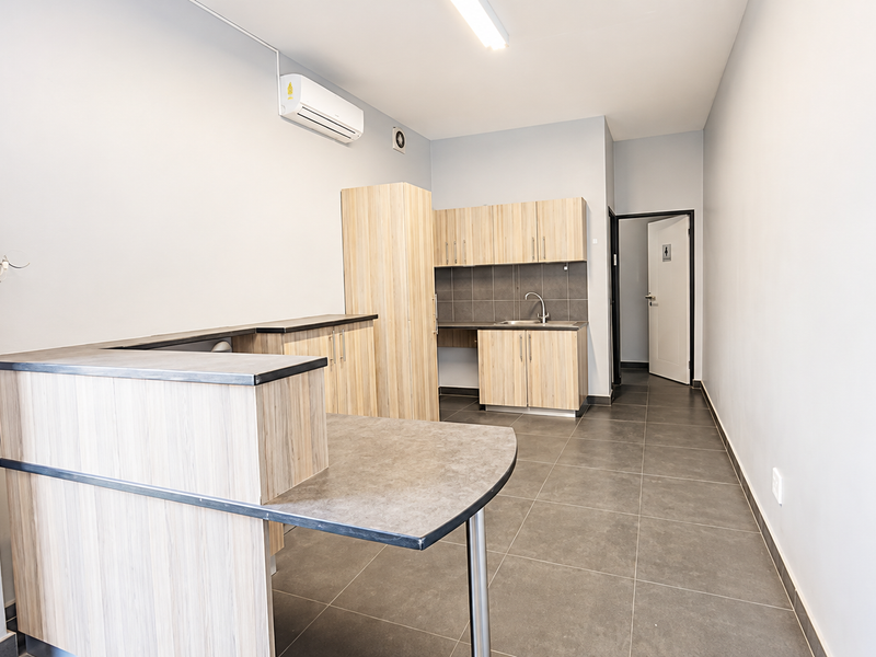 A clean, functional kitchenette area featuring light wood-grain cabinetry, dark laminate countertops, a stainless steel sink, and tiled flooring in an industrial office setting.