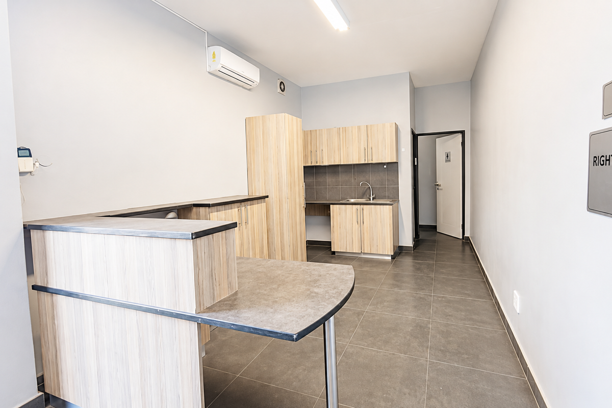 A clean, functional kitchenette area featuring light wood-grain cabinetry, dark laminate countertops, a stainless steel sink, and tiled flooring in an industrial office setting.
