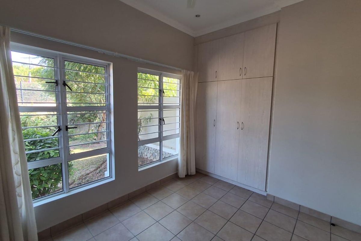A bright, empty bedroom featuring neutral tiled flooring, built-in wooden cupboards, and large windows with security bars overlooking the garden.