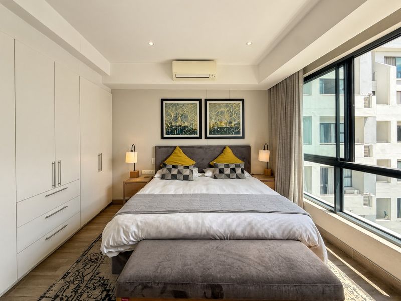 A well-maintained bedroom featuring built-in wardrobes, recessed lighting, a wall-mounted air conditioning unit, and large windows providing natural light.