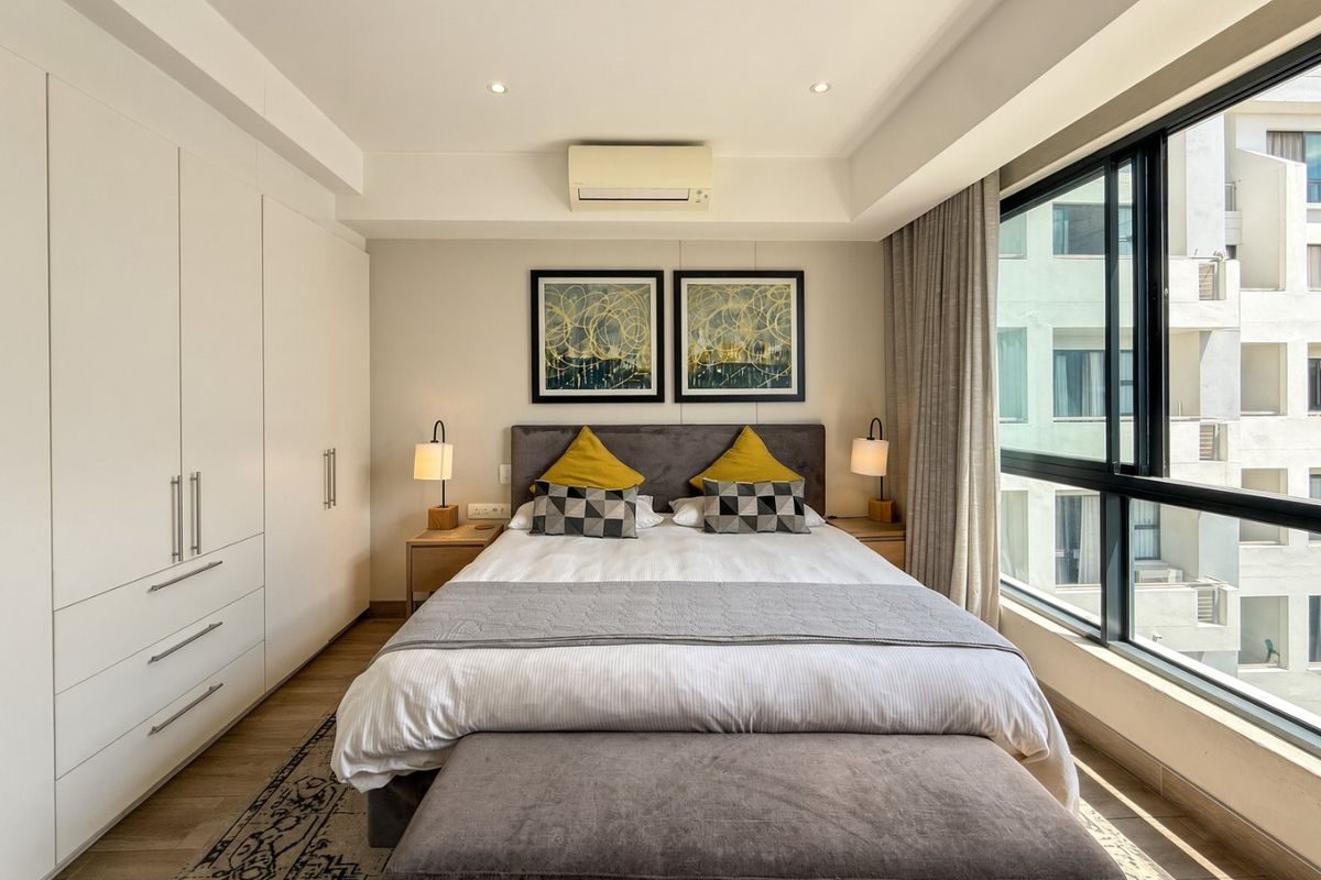 A well-maintained bedroom featuring built-in wardrobes, recessed lighting, a wall-mounted air conditioning unit, and large windows providing natural light.