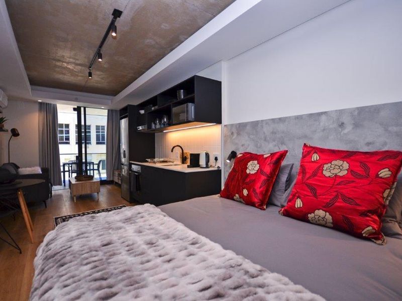 Modern open-plan studio apartment featuring an exposed concrete ceiling, sleek built-in kitchen cabinetry, and direct access to a private balcony.