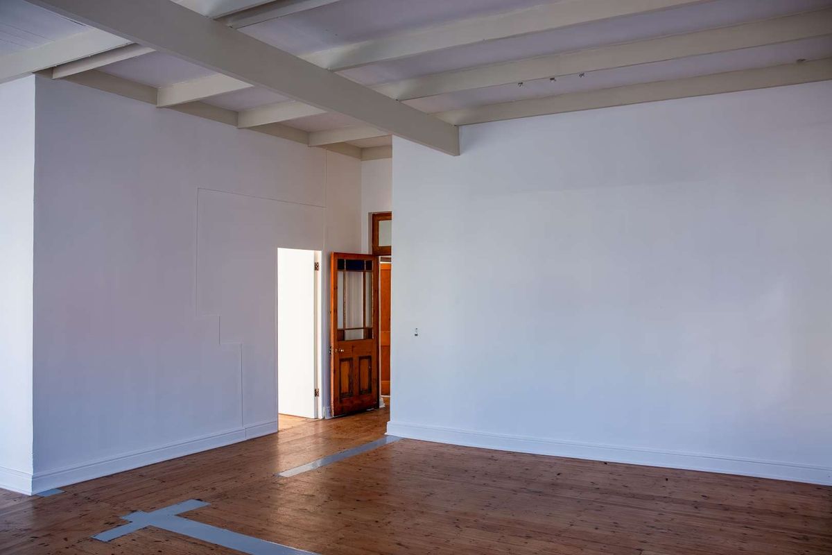 A spacious, empty living area featuring exposed timber ceiling beams and original timber flooring, leading to an adjacent room through a classic timber door.