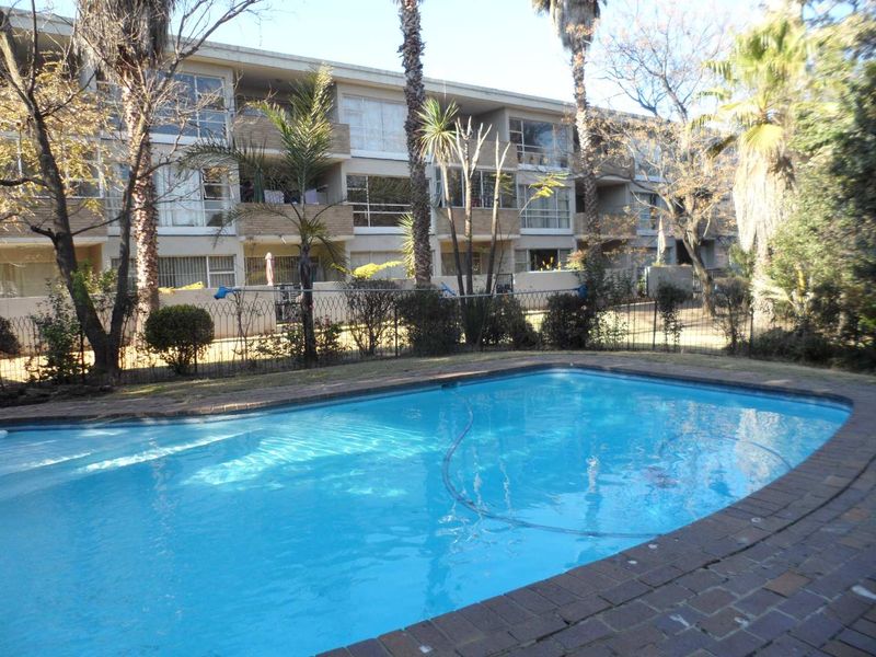 A communal swimming pool area featuring brick coping and metal fencing, set against the backdrop of a multi-story apartment building.