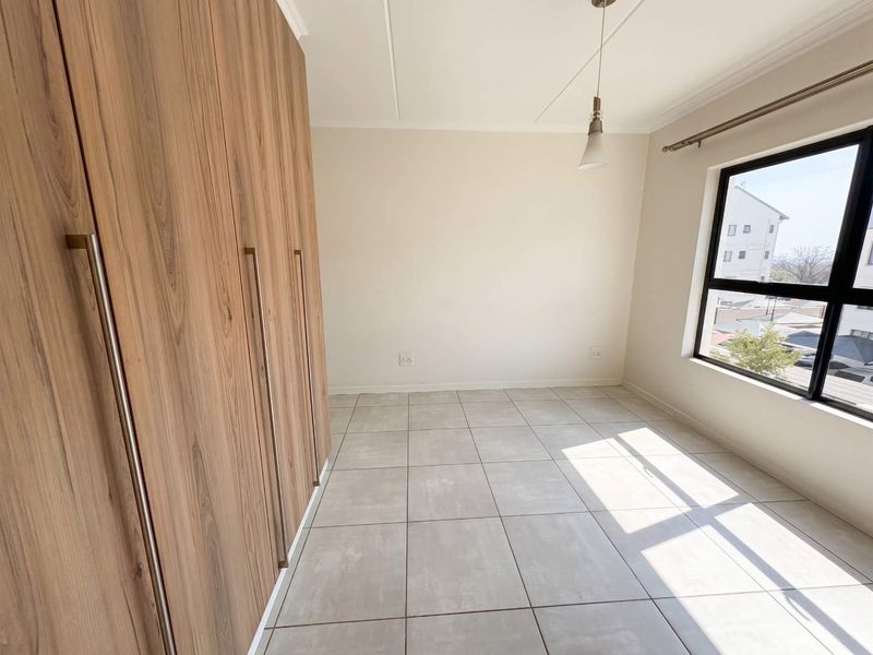 A bright, empty bedroom featuring modern wood-finish built-in cupboards, neutral tiled flooring, and a large window providing natural light.