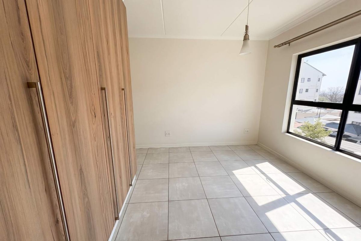 A bright, empty bedroom featuring modern wood-finish built-in cupboards, neutral tiled flooring, and a large window providing natural light.