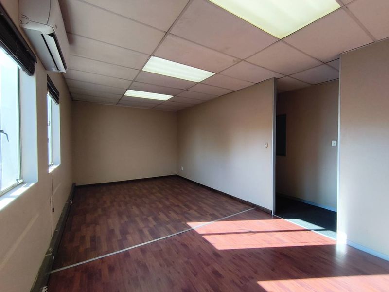 An empty office space featuring laminate wood flooring, a suspended ceiling with recessed lighting, and a wall-mounted air conditioning unit.