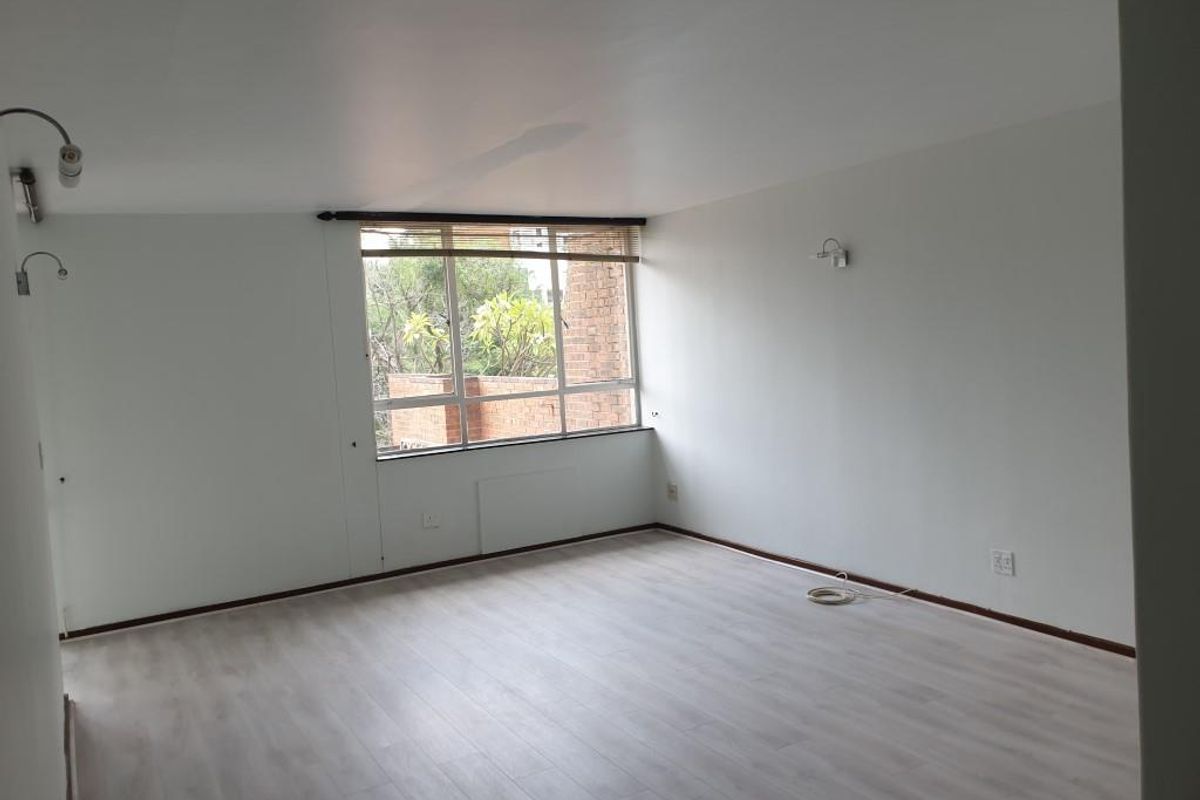 A bright, empty living room featuring light-toned laminate flooring, neutral walls, and a large window providing natural light.