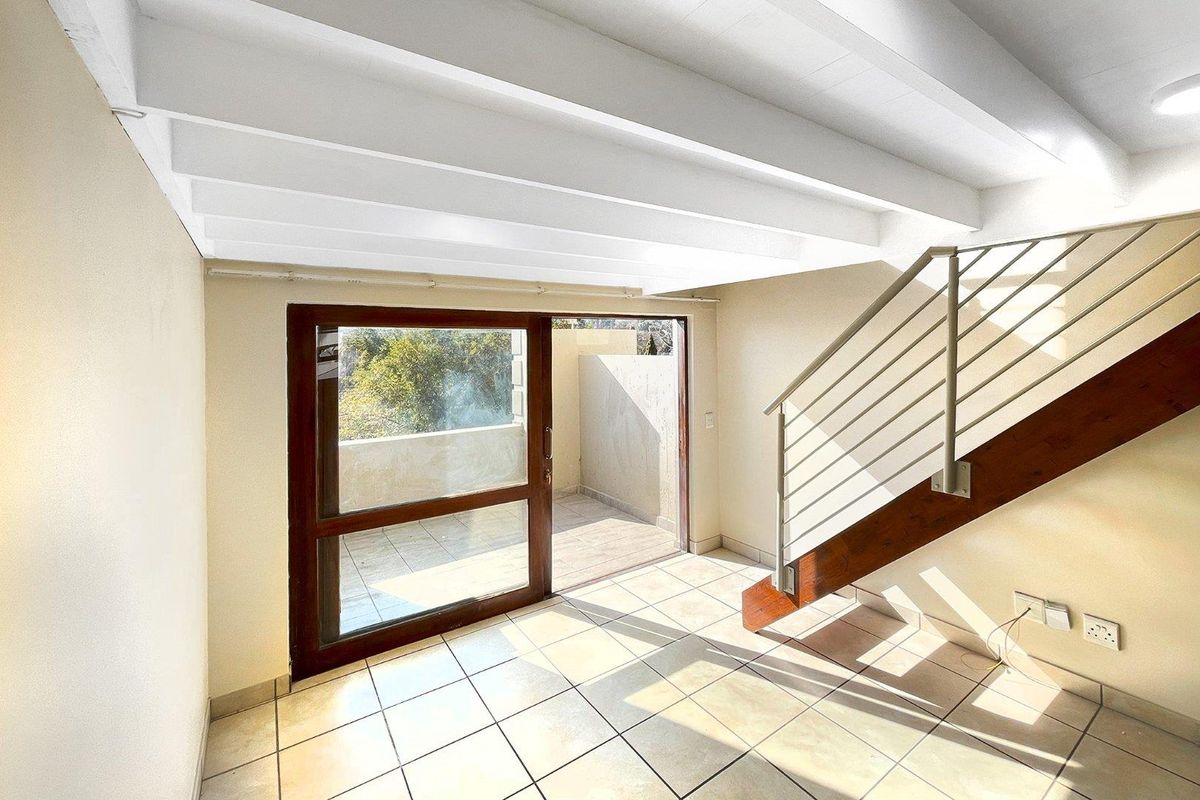 Bright living area featuring exposed white ceiling beams, tiled flooring, a staircase with metal railings, and sliding glass doors opening to a private balcony.