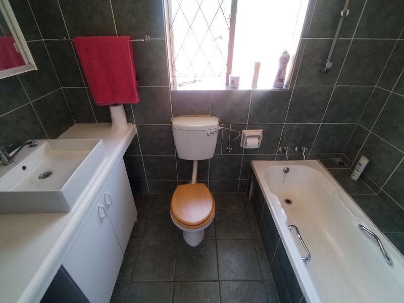 A functional bathroom featuring dark tiled walls and flooring, a white vanity with a vessel sink, a standard toilet, and a built-in bathtub.