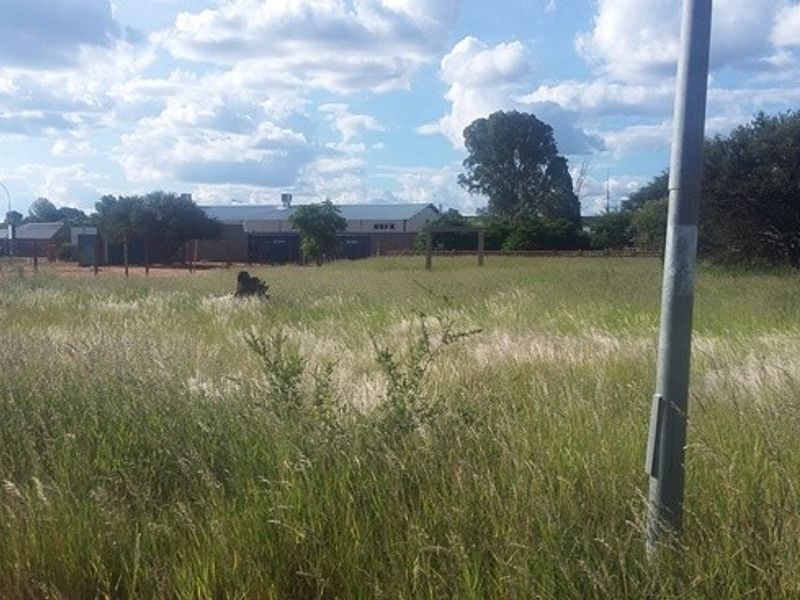 An undeveloped plot of land featuring tall grass, wire fencing, and a utility pole, with distant commercial buildings visible in the background.