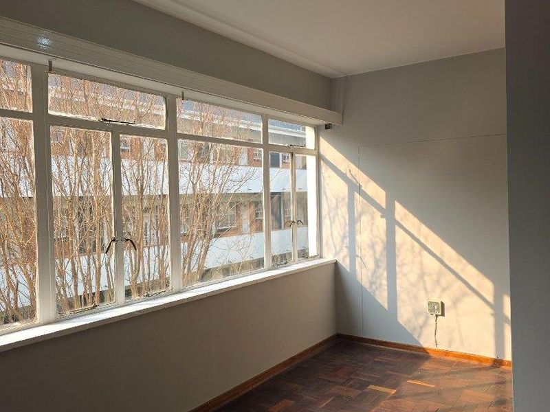 A bright, empty living area featuring classic parquet flooring, large windows providing natural light, and modern track lighting.