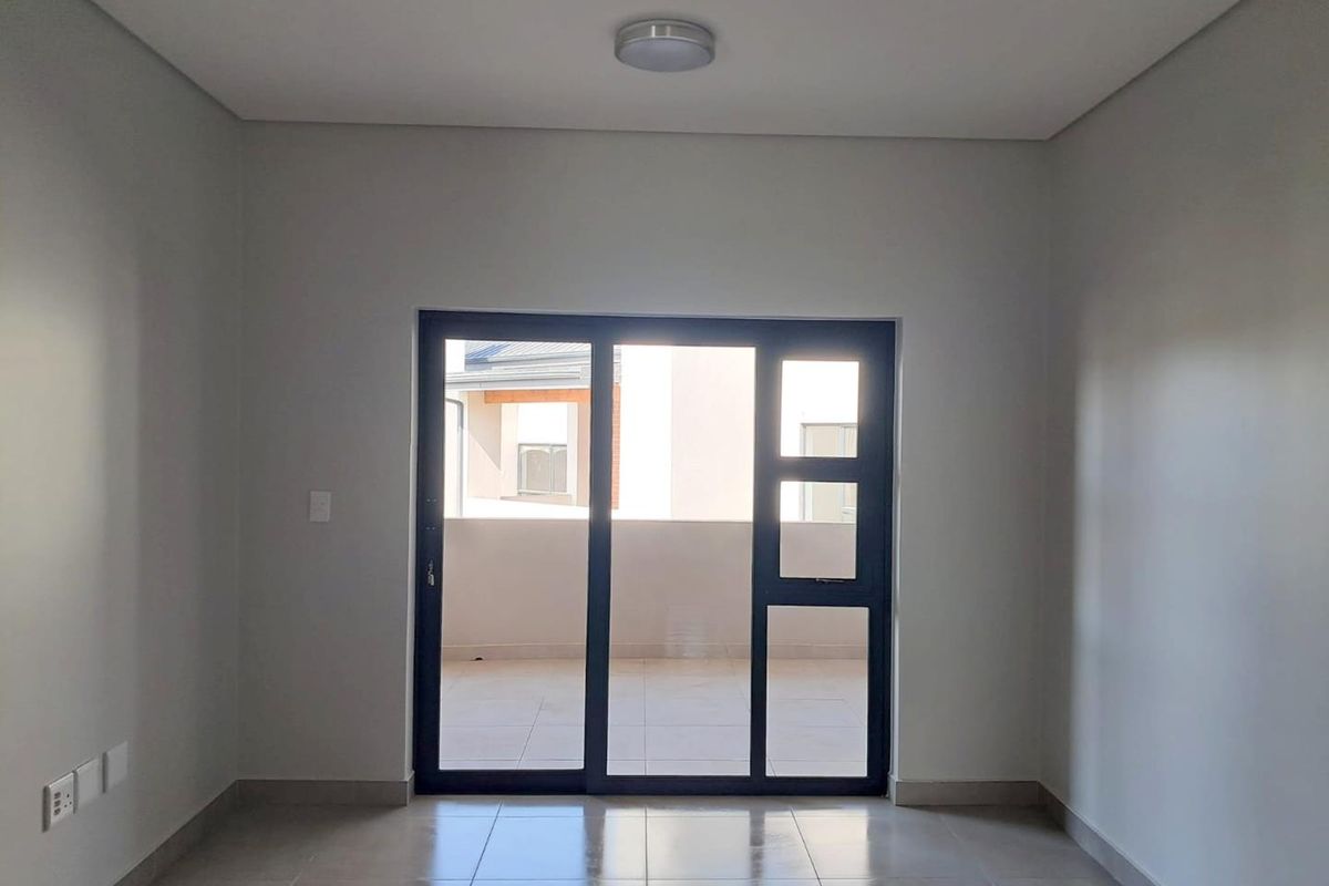 An empty, well-lit living area featuring neutral tiled flooring and a large sliding glass door providing access to an outdoor balcony.
