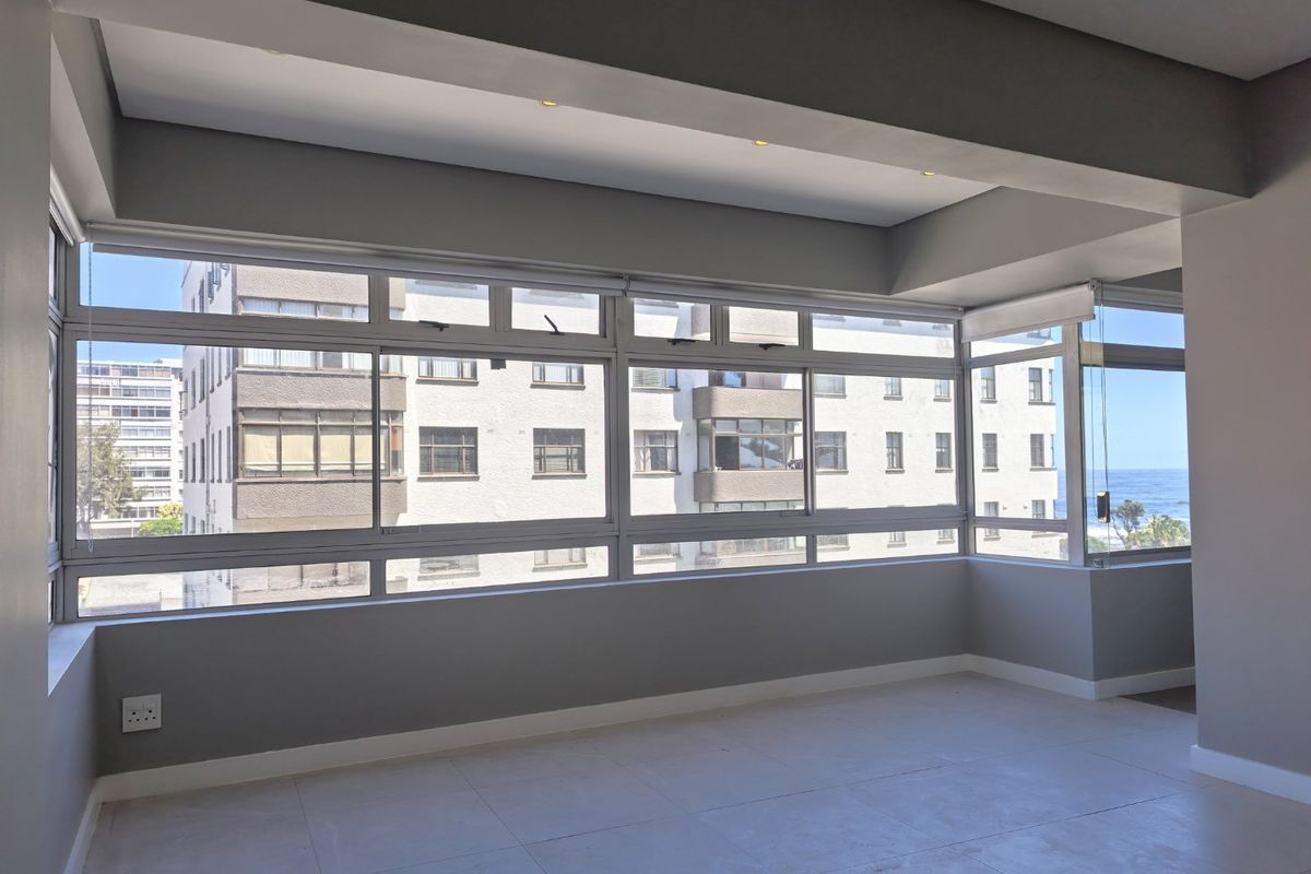 A bright, empty living area featuring large aluminium-framed windows with roller blinds, recessed ceiling lighting, and neutral tiled flooring.