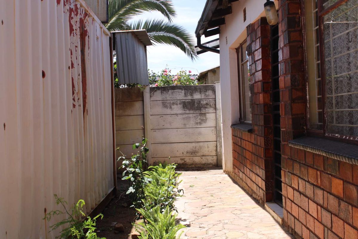 A narrow side passage featuring a paved walkway, brick exterior wall with security bars, and a weathered metal structure.