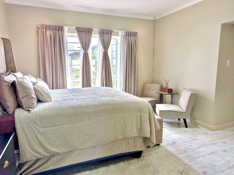 A well-maintained bedroom featuring light-toned laminate flooring, neutral wall finishes, and natural light through a large window with fitted curtains.