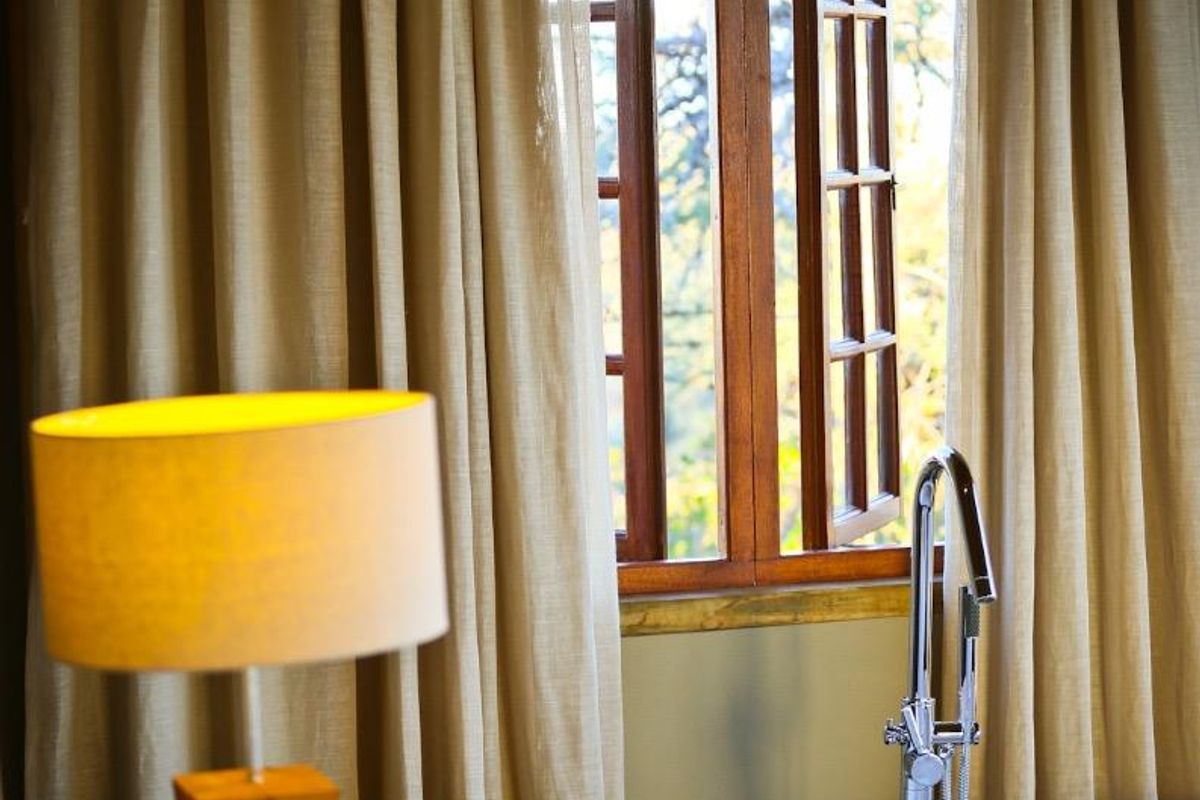 A serene bathroom featuring a freestanding bathtub positioned near a wooden-framed window with natural light and elegant curtains.