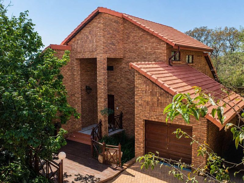 A well-maintained face-brick house featuring a tiled roof, timber decking entrance, and an integrated garage, set within a lush, established garden.