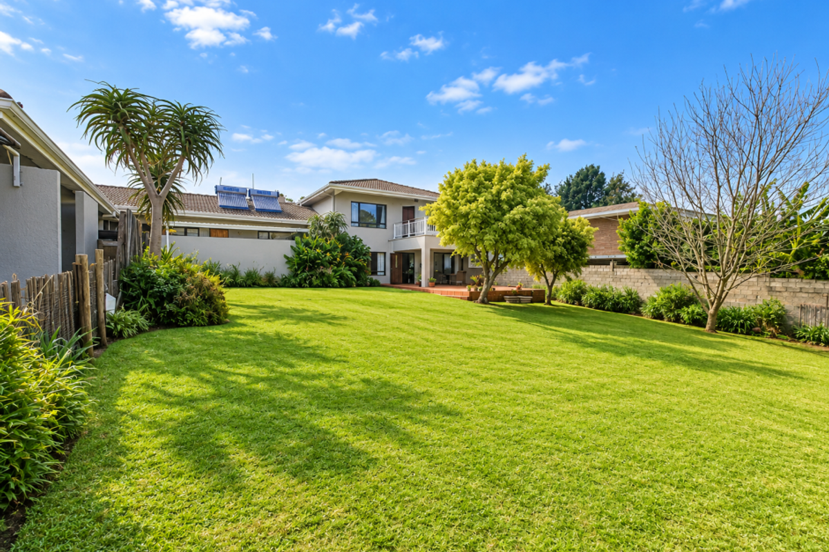 Spacious rear garden featuring a well-maintained lawn, mature trees, and a view of the two-story house exterior with a balcony and solar water heating system.