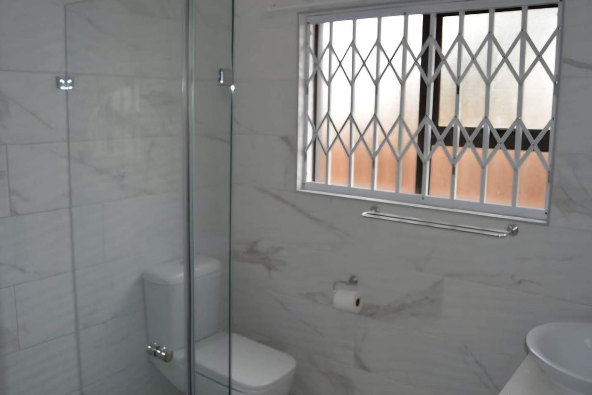 Modern bathroom featuring marble-look wall tiles, a glass-enclosed shower, a wall-mounted toilet, and a vanity basin with a security gate on the window.