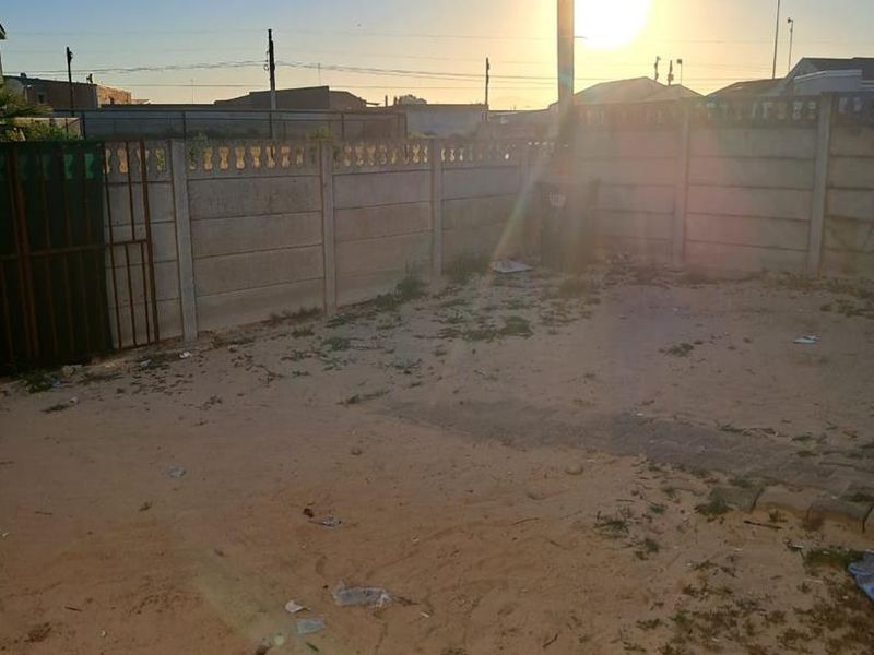 A rear outdoor area featuring a precast concrete boundary wall and metal gate, currently consisting of unlandscaped sandy ground.