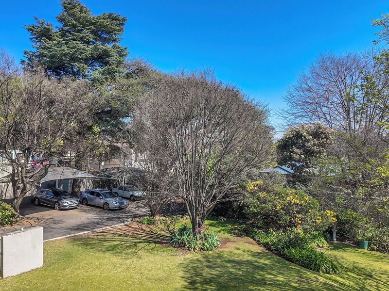An elevated view of the rear property grounds featuring a paved driveway, covered carport parking, and a well-maintained landscaped garden with mature trees.