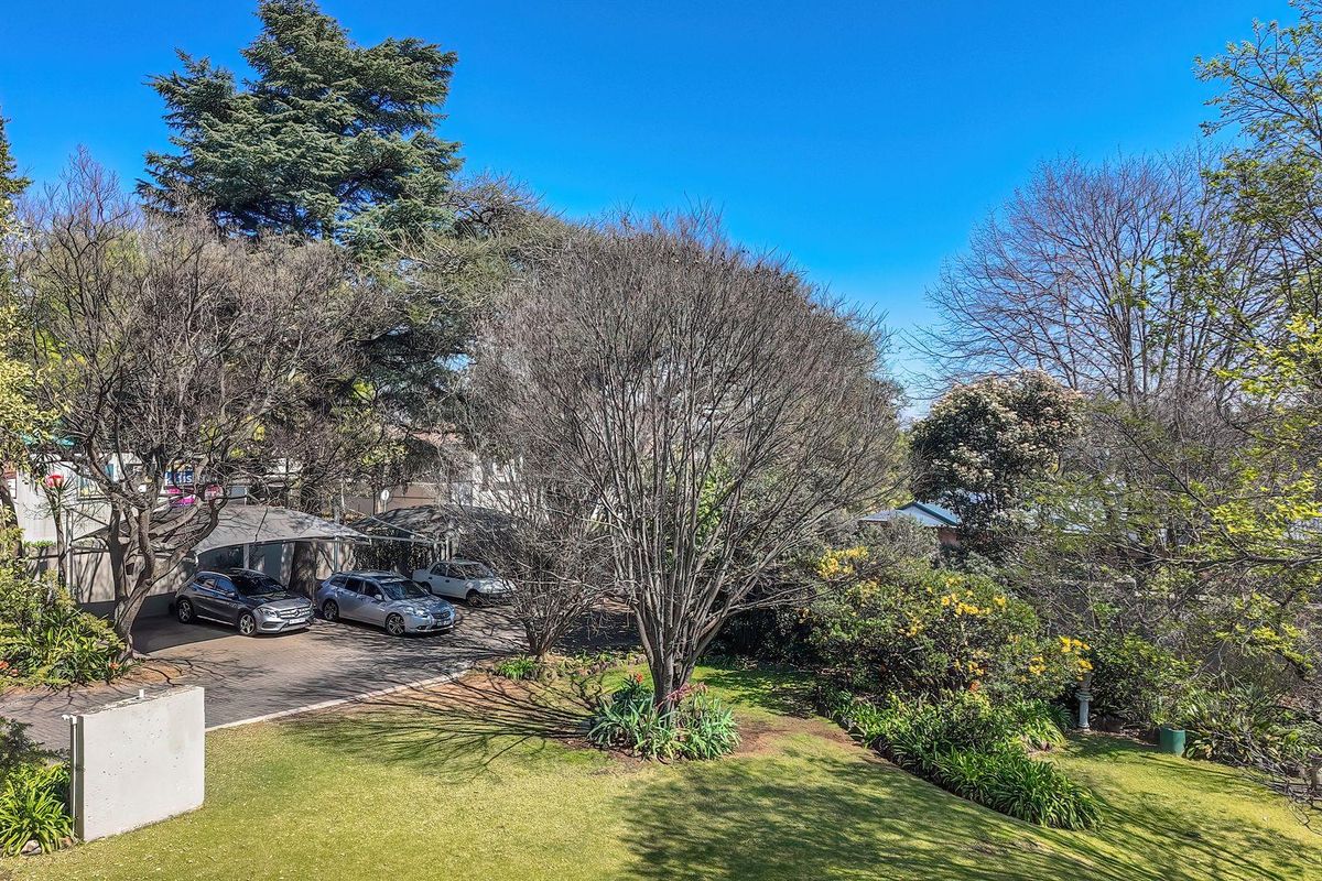 An elevated view of the rear property grounds featuring a paved driveway, covered carport parking, and a well-maintained landscaped garden with mature trees.
